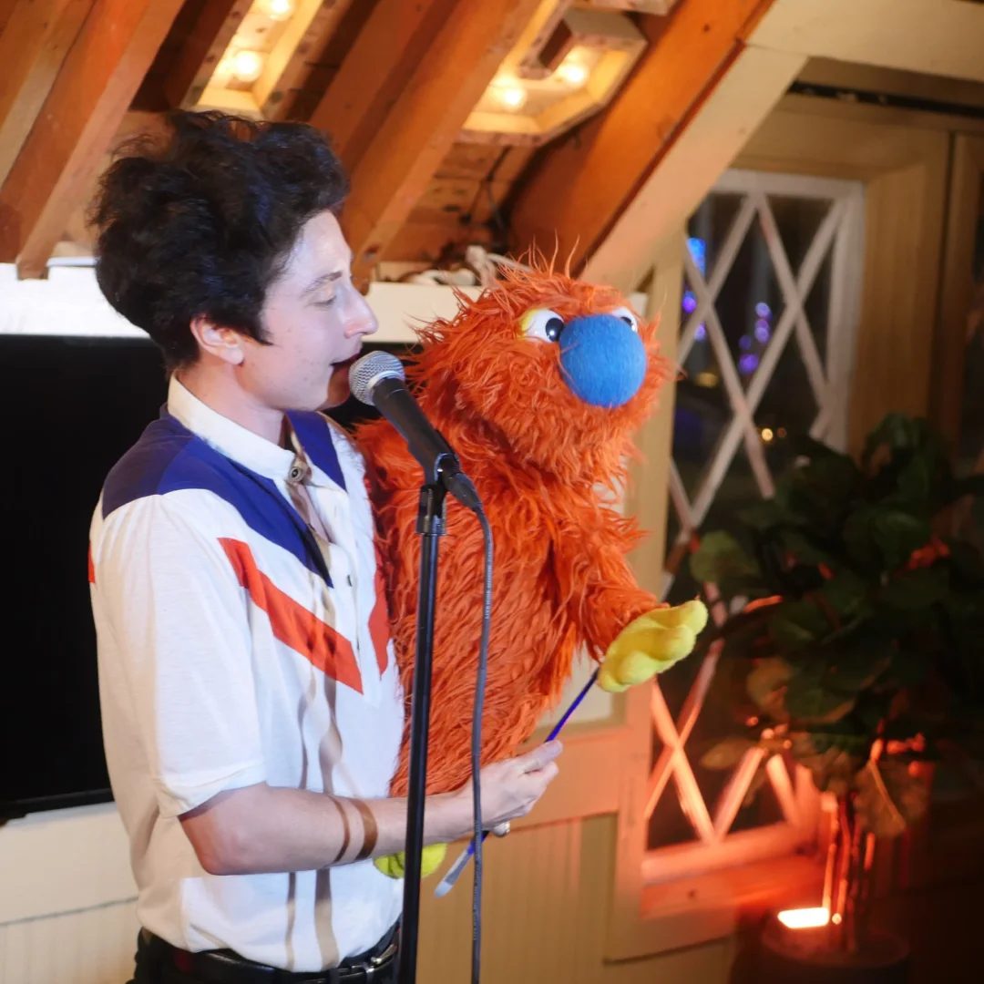 Zack holding his orange puppet Ticklemuff while speaking into a microphone on stage